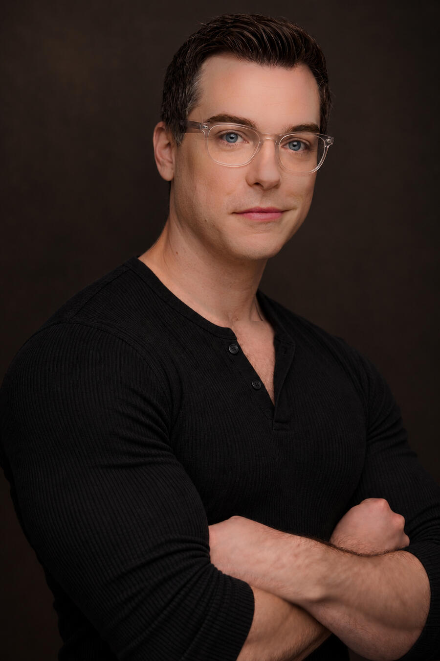 Alec Arden wearing clear glasses and a dark Henley, arms folded against a warm brown background.